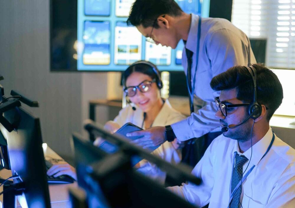 Engineers with headsets working together at a wall of monitoring screens in a dark operations room