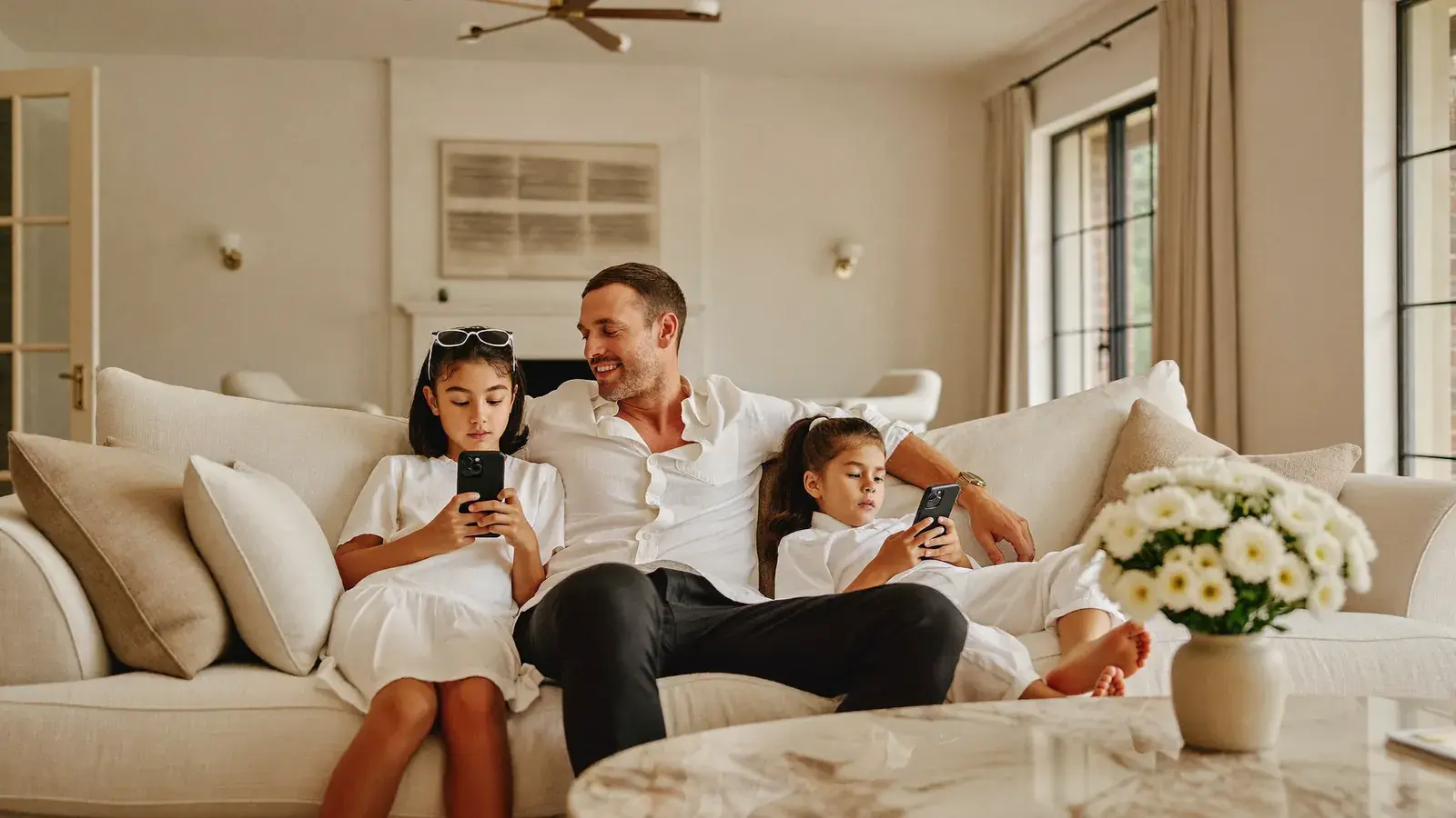 A father relaxes on a cream sofa with his two daughters, each on a phone, in a bright family living room