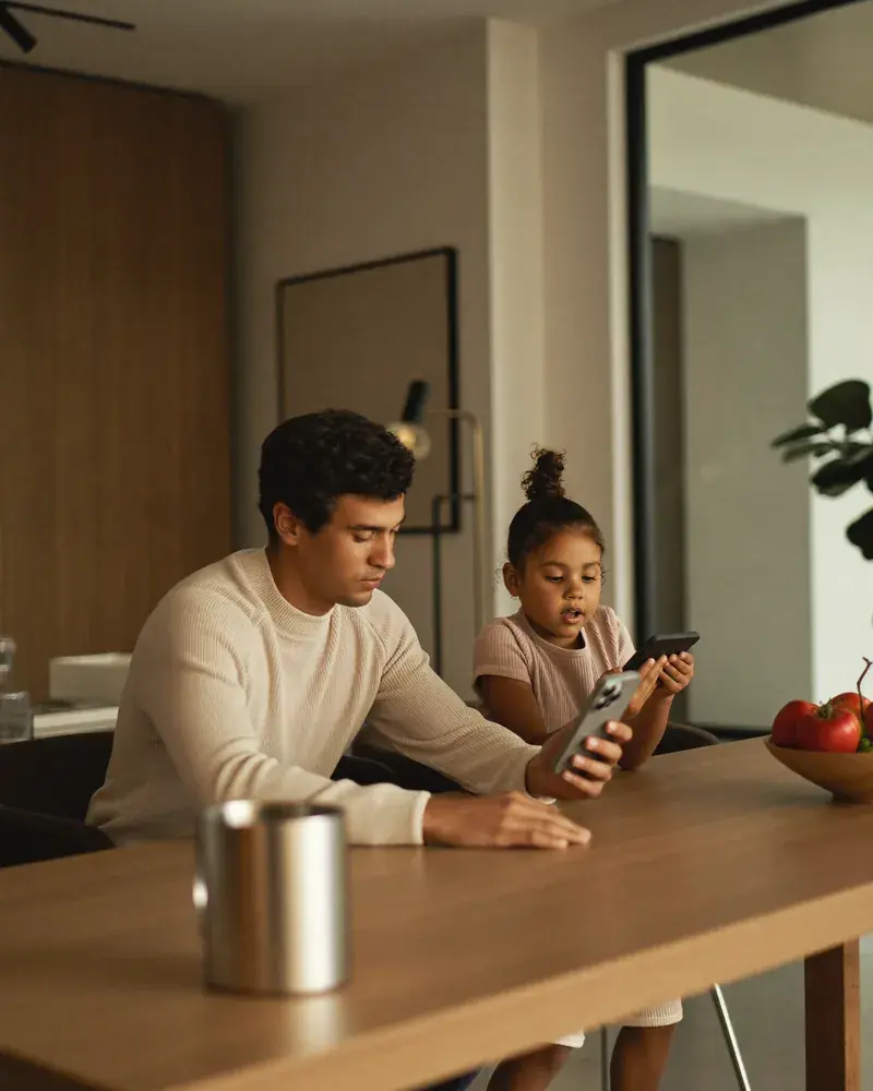 A father and his young daughter check their phones together at the kitchen table of a modern home